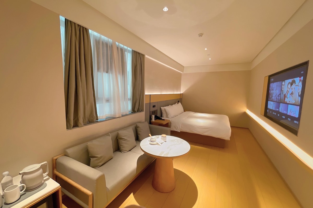 A hotel room with a sofa, a bed, a bedside table with a lamp, a TV on the wall, and a coffee table.
