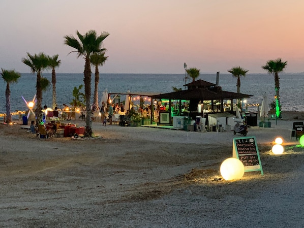 A beachside restaurant with outdoor seating, palm trees, and a sign indicating mask usage.