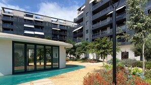 Un edificio moderno con fachada de vidrio, una piscina pequeña y un jardín ajardinado con flores coloridas.