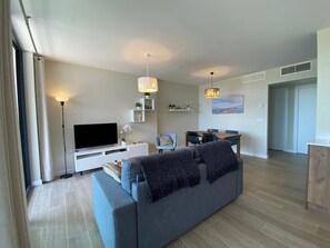 A modern living room with a grey sofa, a flat-screen TV, a dining table with chairs, and pendant lights.