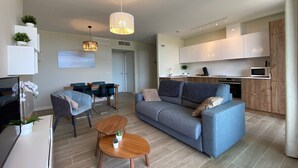 Una sala de estar moderna con sofá, mesa de centro y comedor. Se observa una cocina con armarios y electrodomésticos.