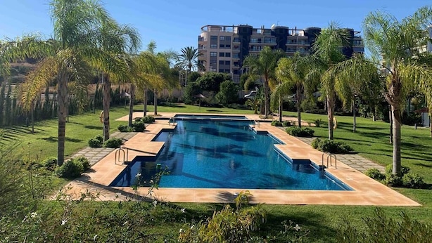 Una piscina rectangular con superficie de agua azul cristalina, rodeada por un césped cuidado y palmeras.
