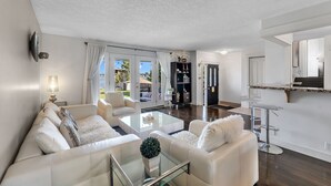 A modern living room with a glass coffee table, white sofas, and a view through large sliding glass doors to an outdoor area.