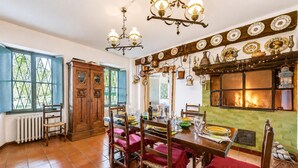 Une salle à manger avec une table en bois dressée pour quatre personnes, entourée de chaises. On peut voir une cheminée avec des pots suspendus et un mur recouvert de carreaux verts.