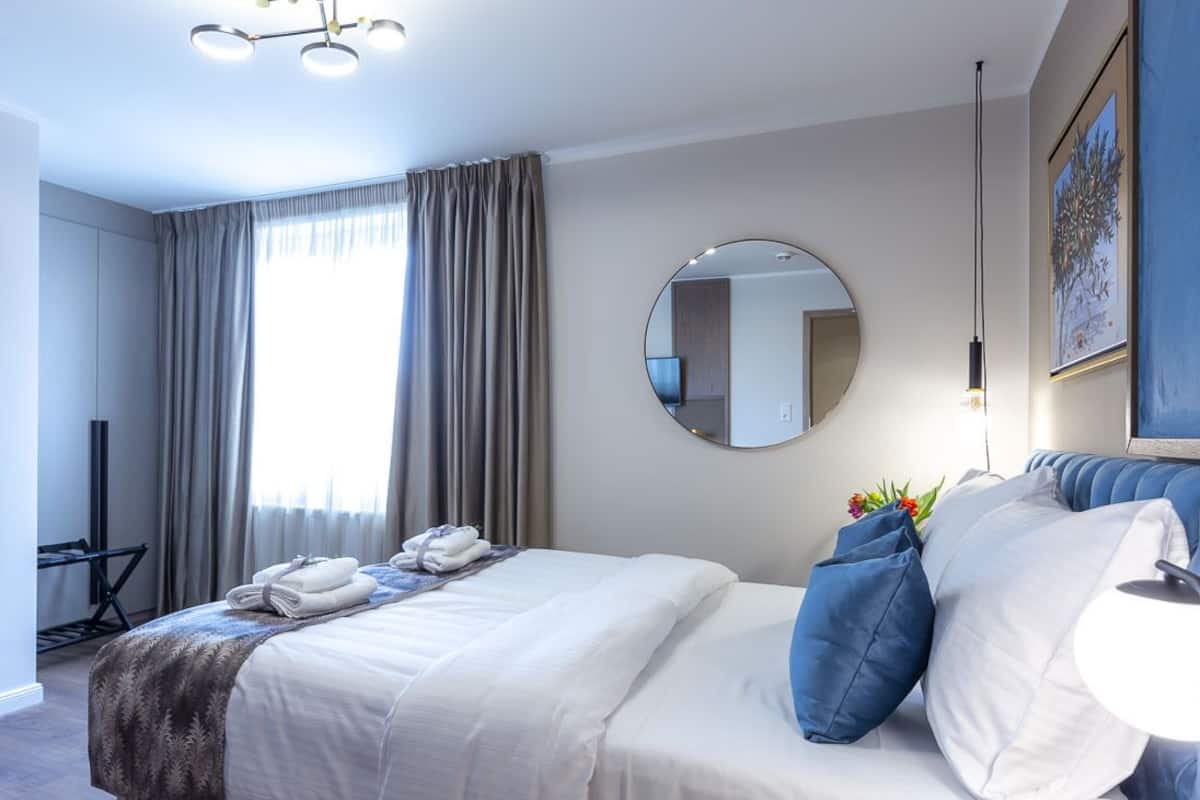 A modern hotel room with a neatly made bed, a round mirror, a desk with a chair, and a large window with curtains.