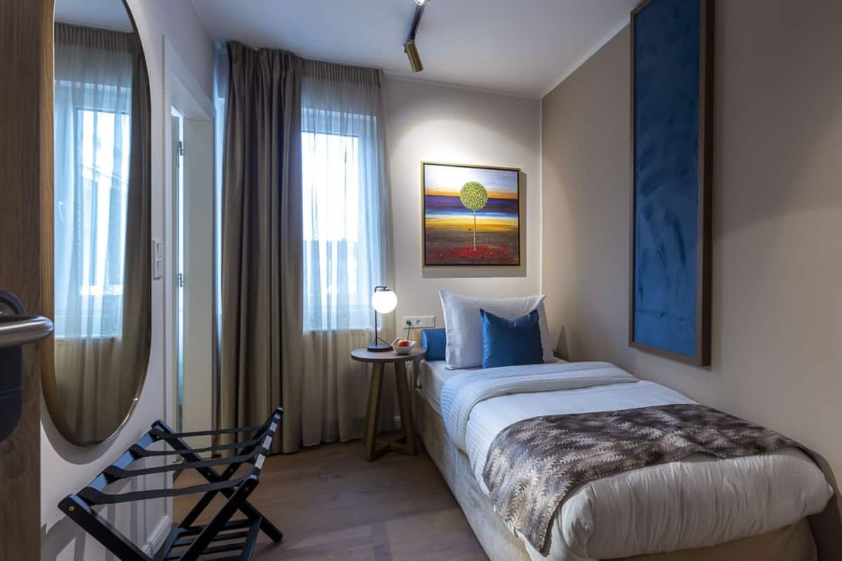 A hotel room with a bed, a small table with a lamp, a chair, and a painting on the wall.
