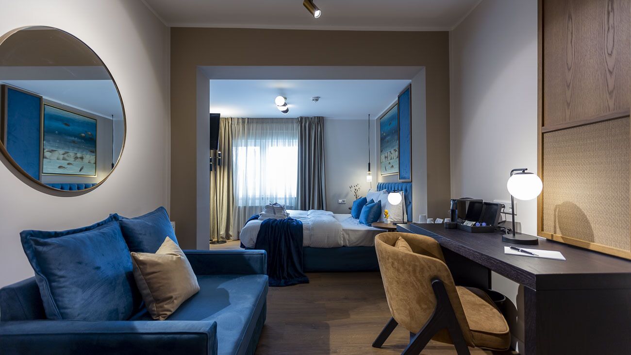 A hotel room with a bed, a desk with a lamp, a chair, a round mirror, and a blue sofa.