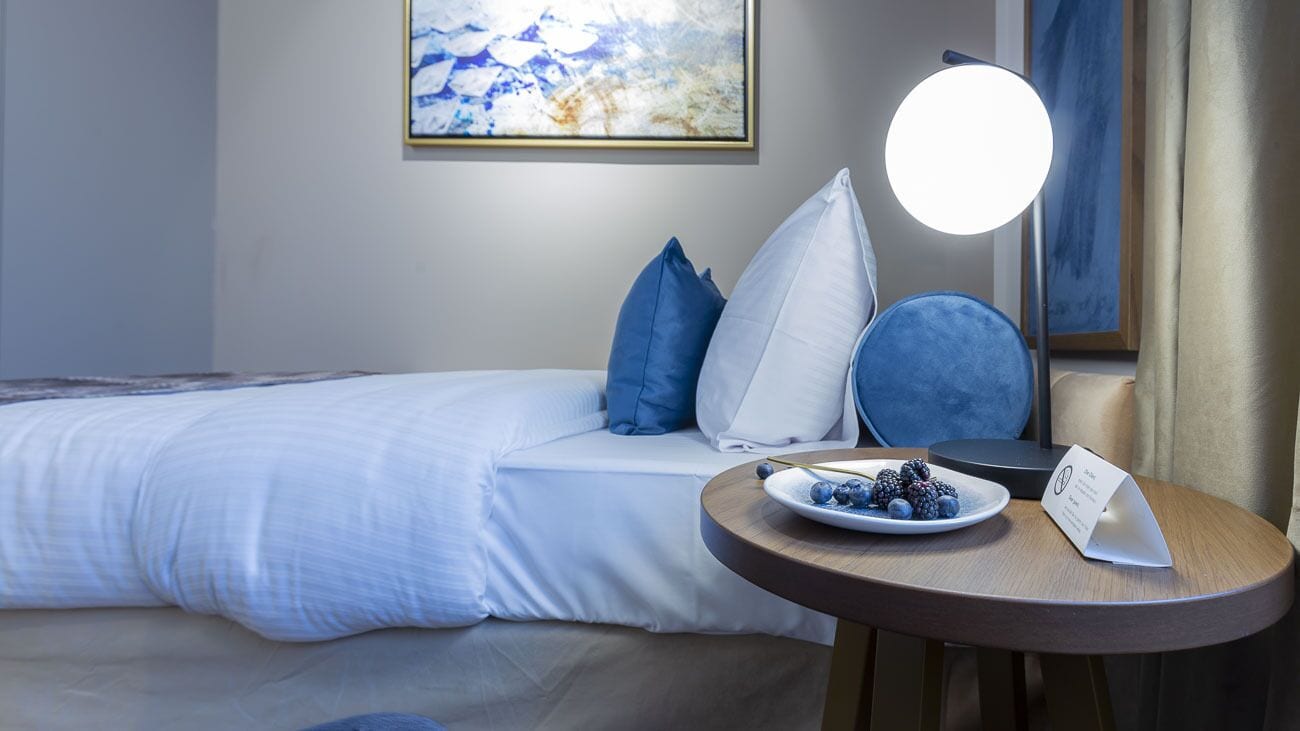 Hotelkamer met een bed, een rond tafel met een schaal bessen, een lamp en een abstract schilderij in een lijst aan de muur.