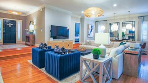 Ein geräumiges Wohnzimmer mit großem Fernseher, blau-weißen Sofas, einem Couchtisch und einem Essbereich mit Lampe und Stühlen.