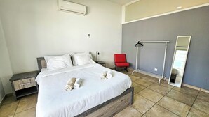 En pænt redt seng med to sammenrullede håndklæder, en rød stol, et vægstående spejl og en fastmonteret aircondition.