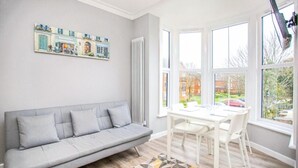 A modern living room with a grey sofa, a dining table with chairs, a radiator, and a large window offering a view of the outside. - Watford WD18 - Homely 2Bedroom/1Bathroom Flat w/ FREE WIFI & FREE PARKING (Watford)