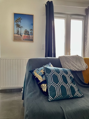 Une pièce avec un lit, un tableau représentant une scène de plage, une fenêtre avec des rideaux et un radiateur.