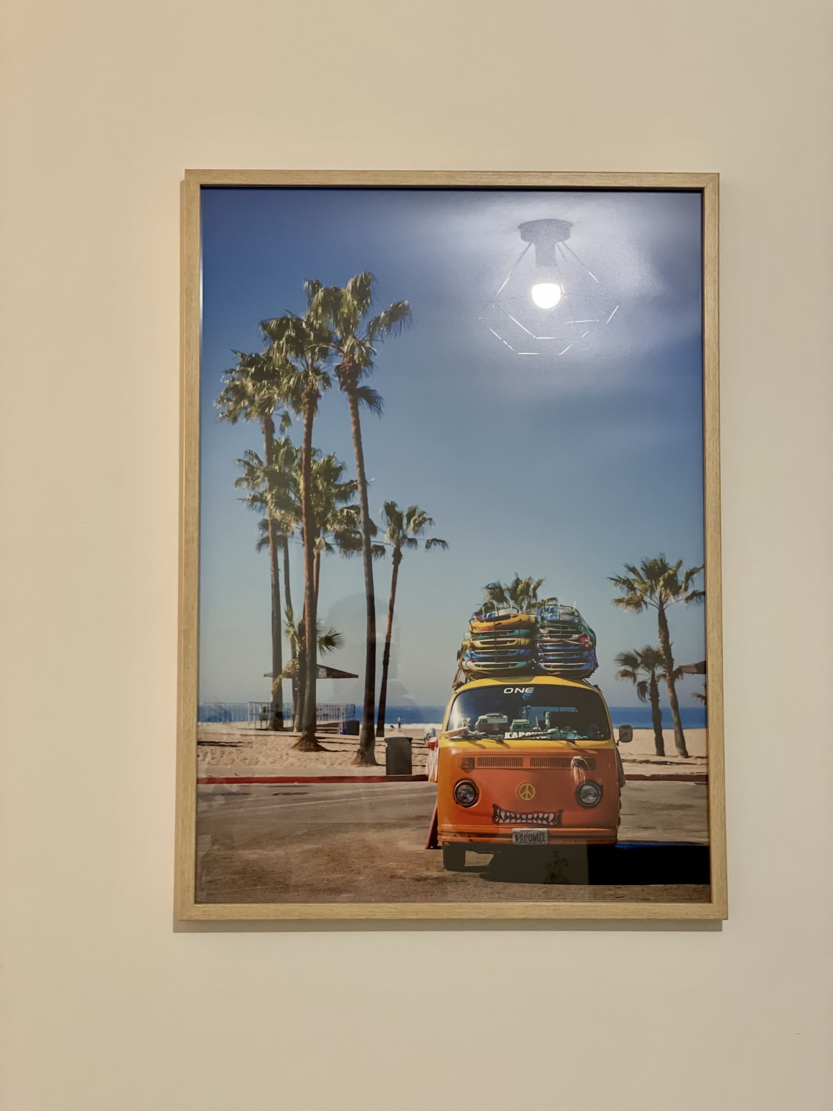 Una obra de arte enmarco muestra una furgoneta con una casa rodante en la parte superior, estacionada en una playa con palmeras y el océano al fondo.