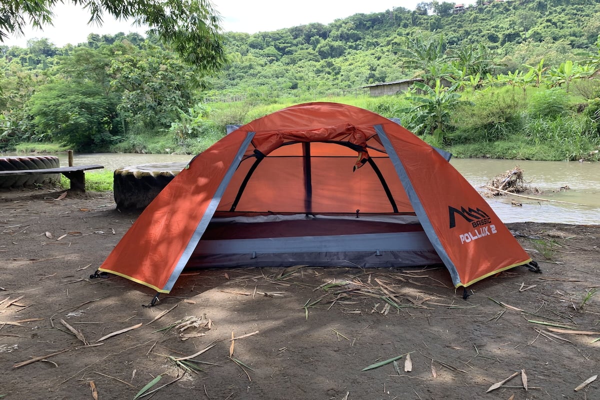 A red tent with a logo, set up on a dirt ground near a river with lush greenery in the background.