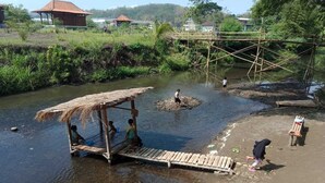 A riverbank with a thatched-roof structure, a wooden bridge, and a small dock. - Camping Ground Taman Nggirli (Srimulyo)