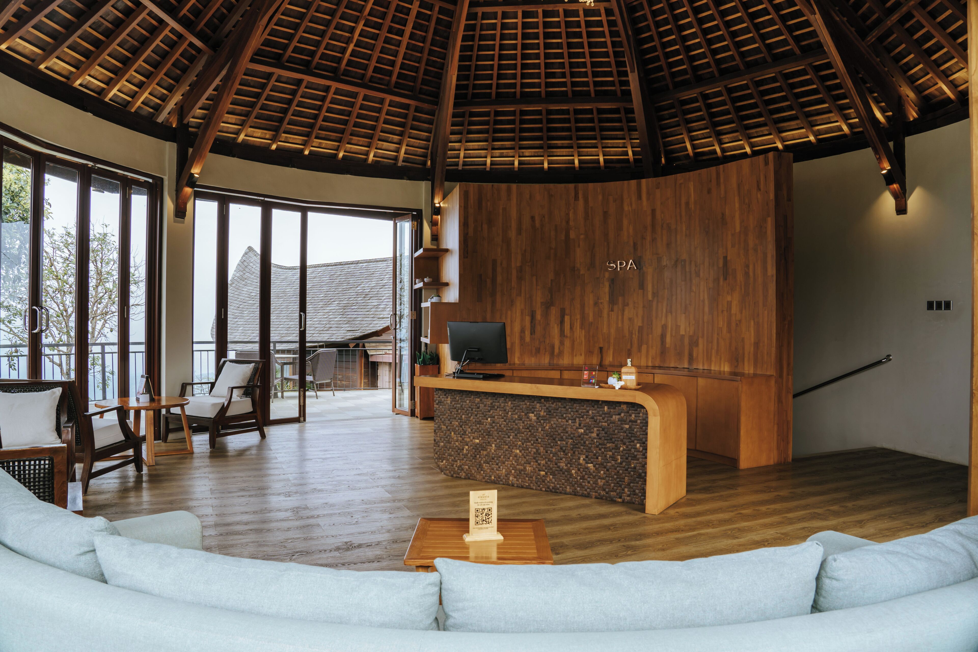 A spacious lobby with a wooden reception desk, seating area, and large windows offering a view of the outside.