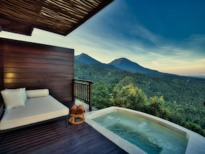 A wooden deck with a sofa and a hot tub overlooking a forested mountain range.