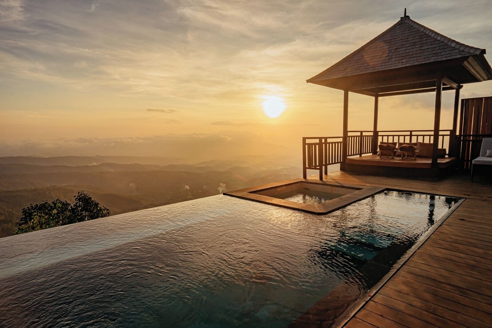 An infinity pool with a wooden deck and a gazebo overlooking a mountainous landscape during sunset.