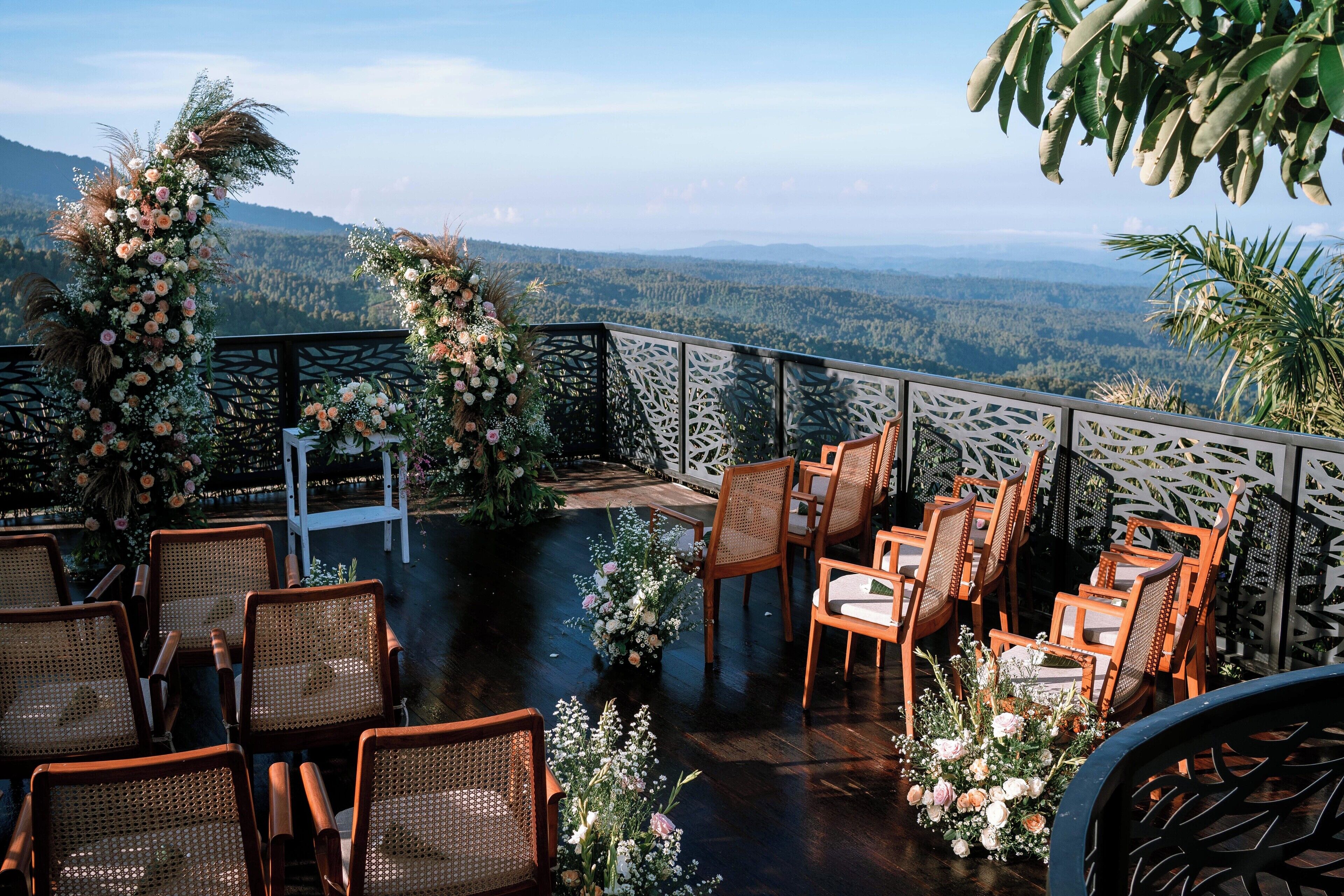 An outdoor seating area with wicker chairs and tables, surrounded by floral arrangements and a view of a mountainous landscape.