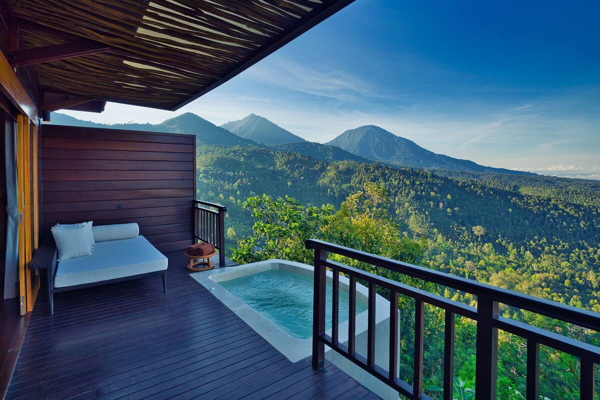 Terraza de madera con un jacuzzi y una cama con vistas a una cadena montañosa boscosa.