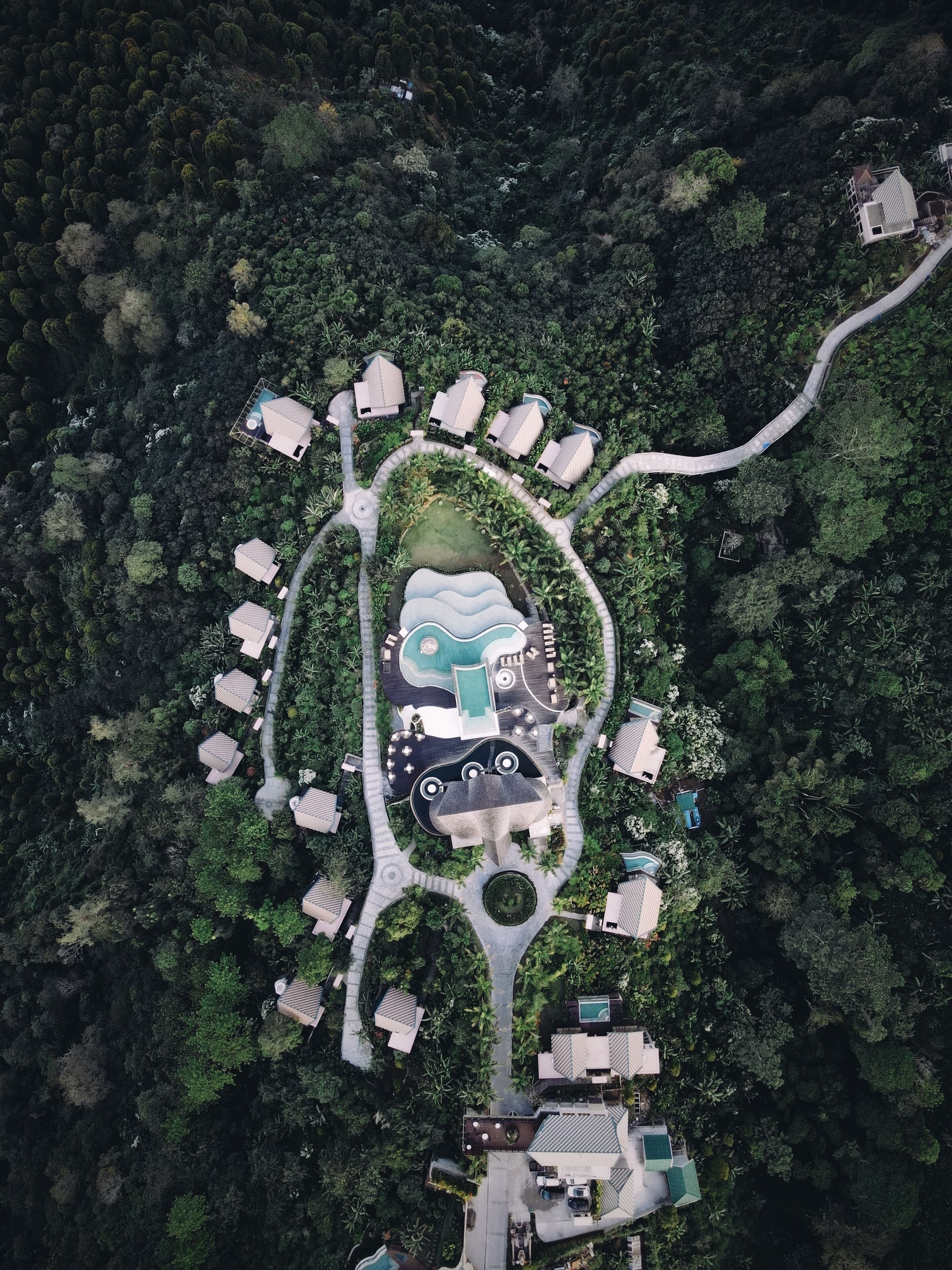Vue aérienne d’un complexe hôtelier comprenant plusieurs hébergements de type bungalow, une piscine centrale et une route sinueuse.