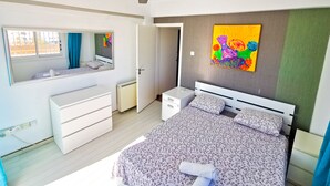 Quarto com cama, cômoda, espelho, ventilador de teto e um quadro colorido na parede.