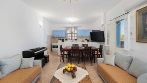 A living room with a piano, sofas, a dining table, and a fruit bowl. - Villa Kocis with Private Pool (Santorini)