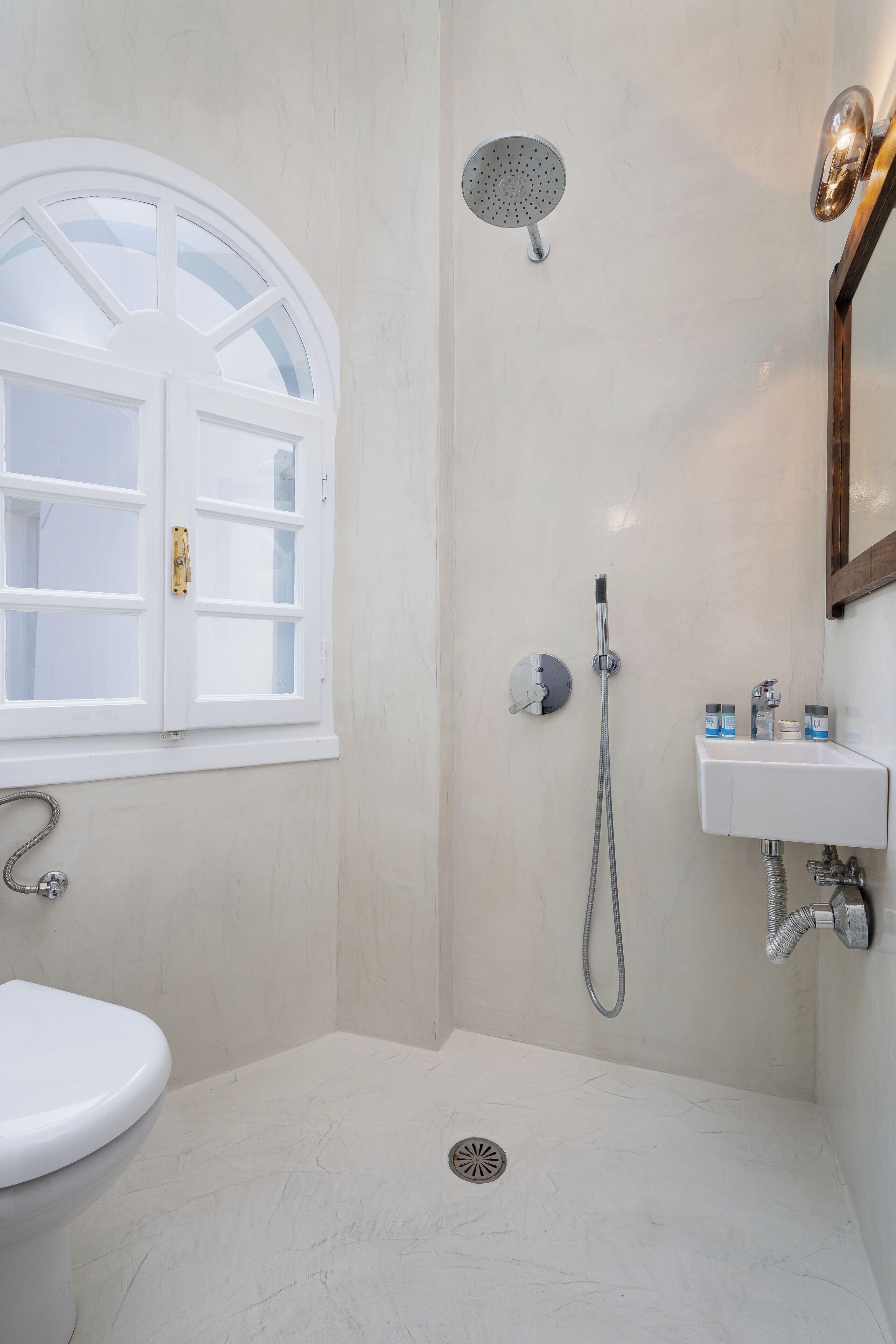 Un baño con inodoro blanco, ducha con cabezal de mano, lavamanos montado en la pared y una ventana con marco blanco.