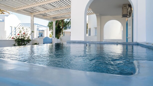 A swimming pool with a white pergola overhead, a covered archway, and a view of a white building with blue accents. - Villa Kocis with Private Pool (Santorini)