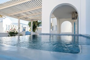 Una piscina con un pergolado blanco sobre ella, un arco cubierto y vistas de un edificio blanco con detalles azules.