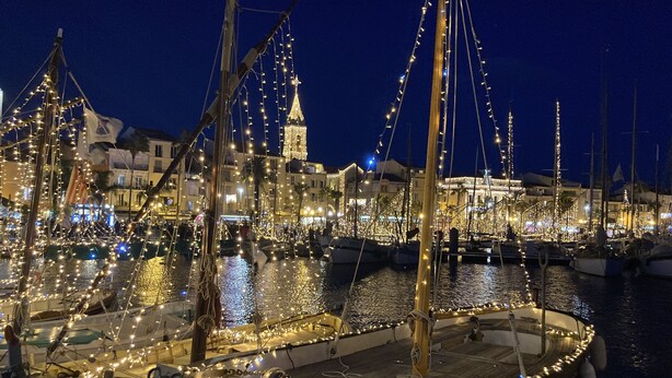 A marina at night with boats decorated with lights, a prominent tower, and buildings in the background.