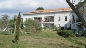 A two-story building with a tiled roof, surrounded by a grassy area with trees and shrubs.