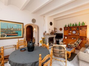 Een gezellige woonkamer met een ronde tafel, houten stoelen, een open haard en een boekenplank vol flessen.