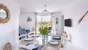 A dining area with a round table set for four, white chairs, a vase with flowers, and a view of the outdoors through a glass door. - La Perle de Damgan by Interhome (Damgan)