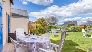 A patio with a table set for dining, surrounded by white chairs, and a garden with various plants and a fence in the background. - La Perle de Damgan by Interhome (Damgan)