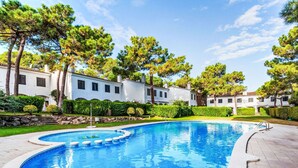 Una piscina con borde curvo, rodeada por una zona pavimentada. Se ven árboles y un edificio con fachada blanca al fondo.