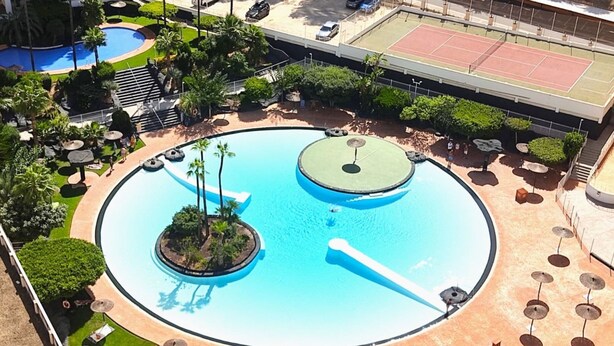 Una piscina circular con dos islotes, rodeada de palmeras y tumbonas.