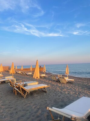 A beach with rows of sun loungers and umbrellas, a sandy shore, and the ocean in the background.