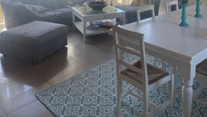 En stue med sofa, skammel, sofabord og et spisebord med stole. Gardiner ved vinduerne, og en radiator er synlig.