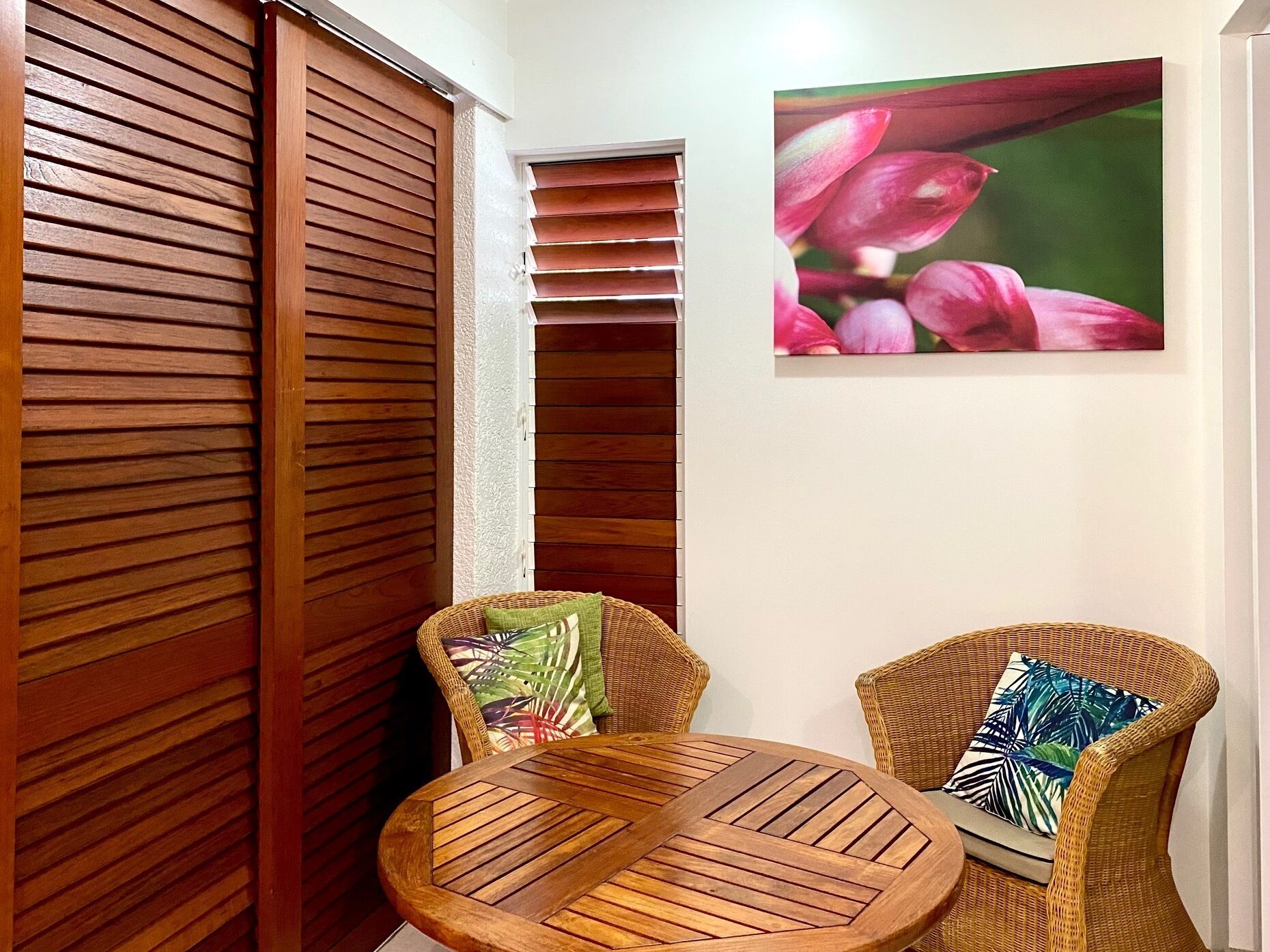 A room with a wooden sliding door, a round wooden table, two wicker chairs, and a wall-mounted picture of pink flowers.