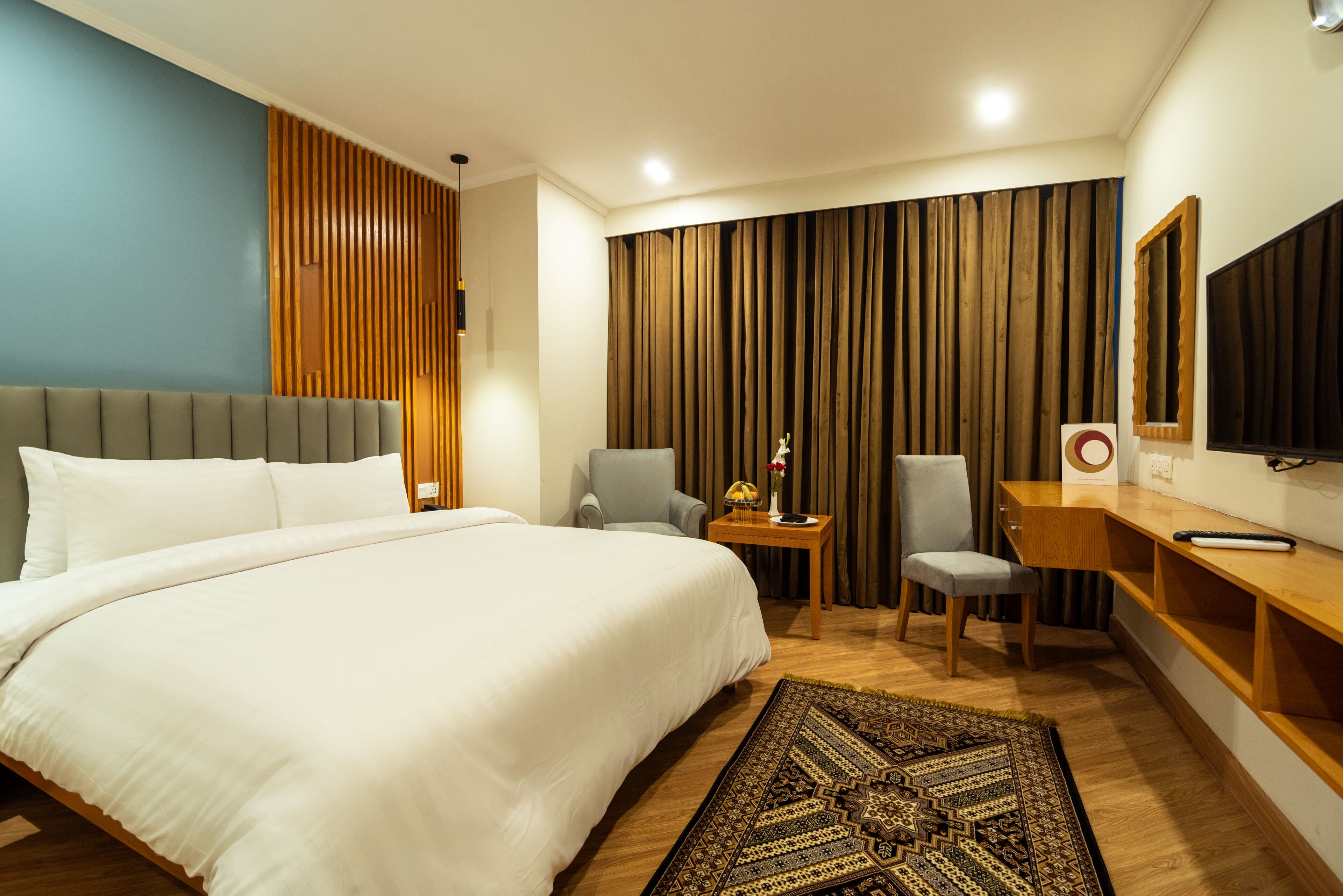 A hotel room with a large bed, two chairs, a desk, a TV, and a patterned rug.