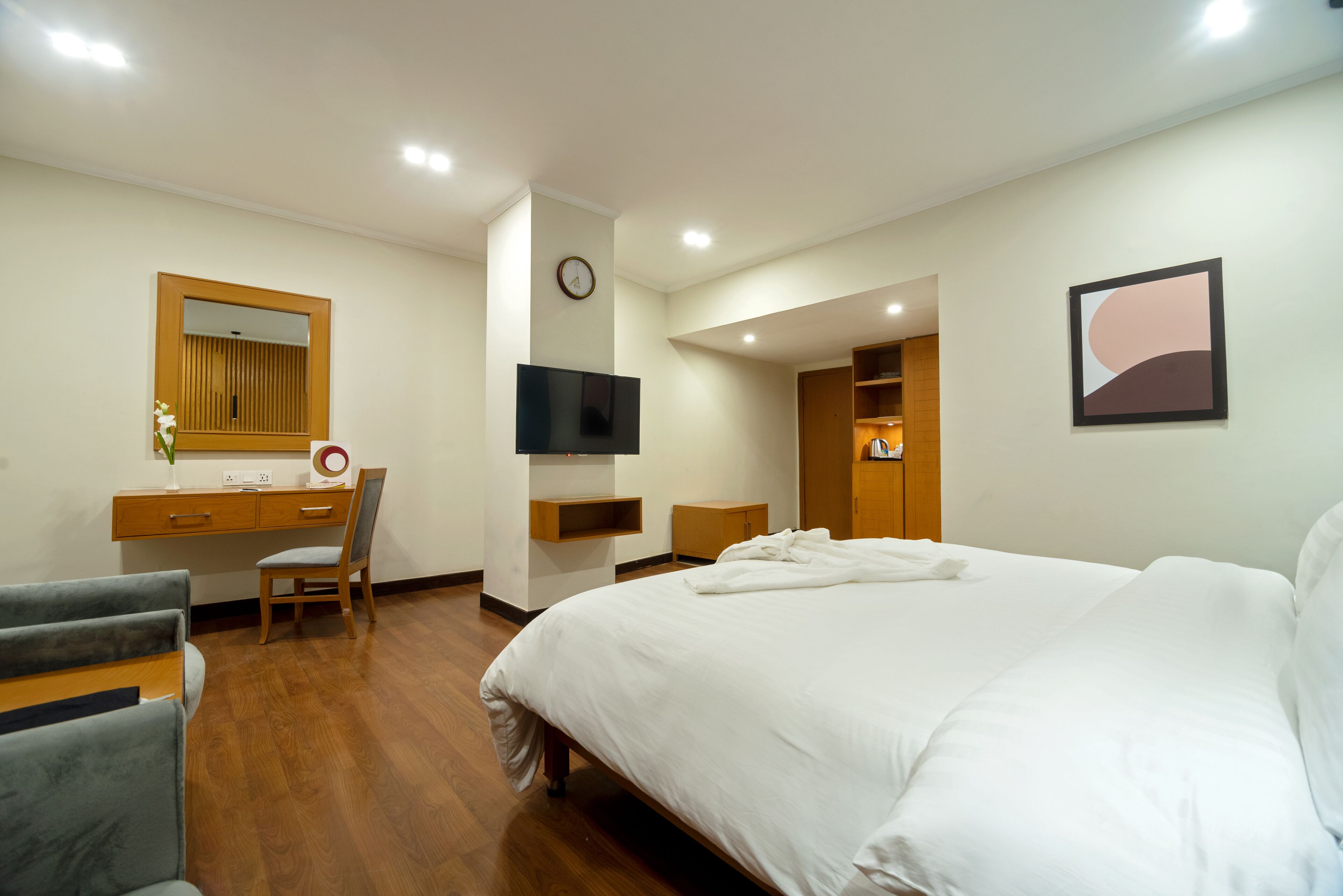 A hotel room with a bed, a desk, a chair, a TV, a clock, and a painting on the wall.