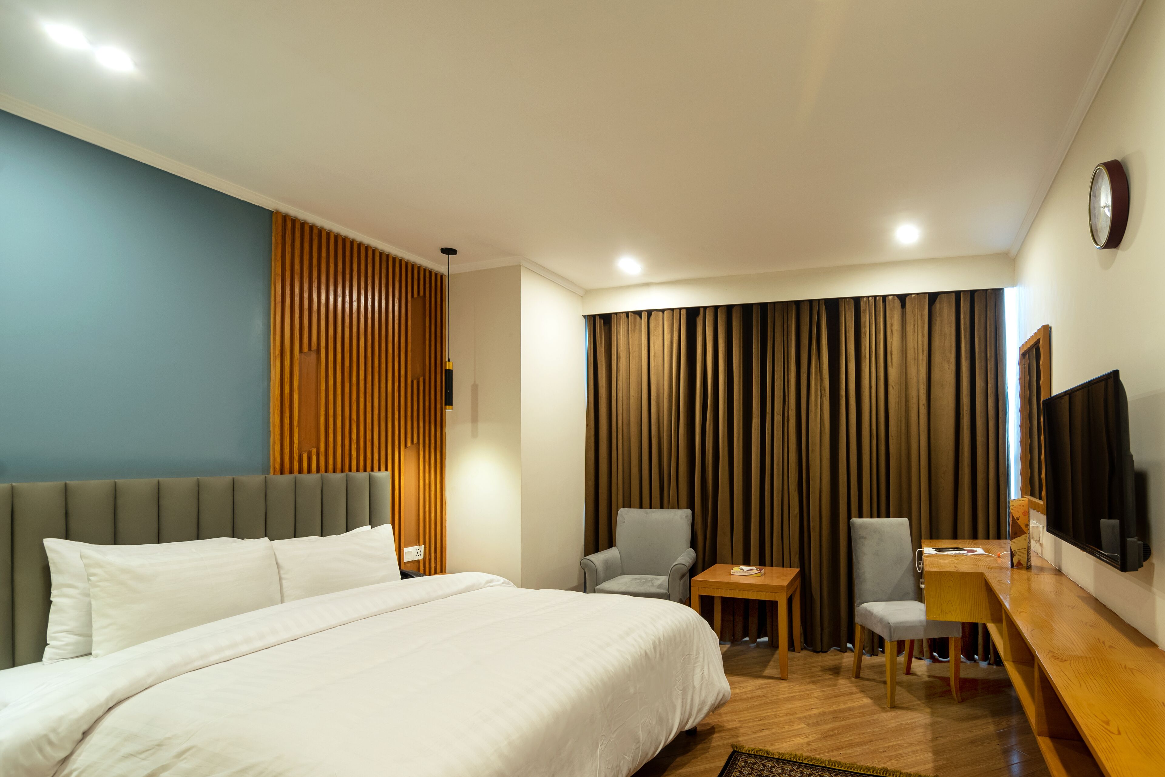 Quarto de hotel com uma cama grande, duas cadeiras, uma mesinha, uma TV e uma cortina.