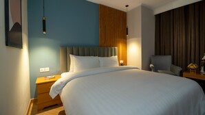 En snyggt bäddad säng i ett modernt hotellrum, med ett träbord vid sängen, en stol och en vägglampa.