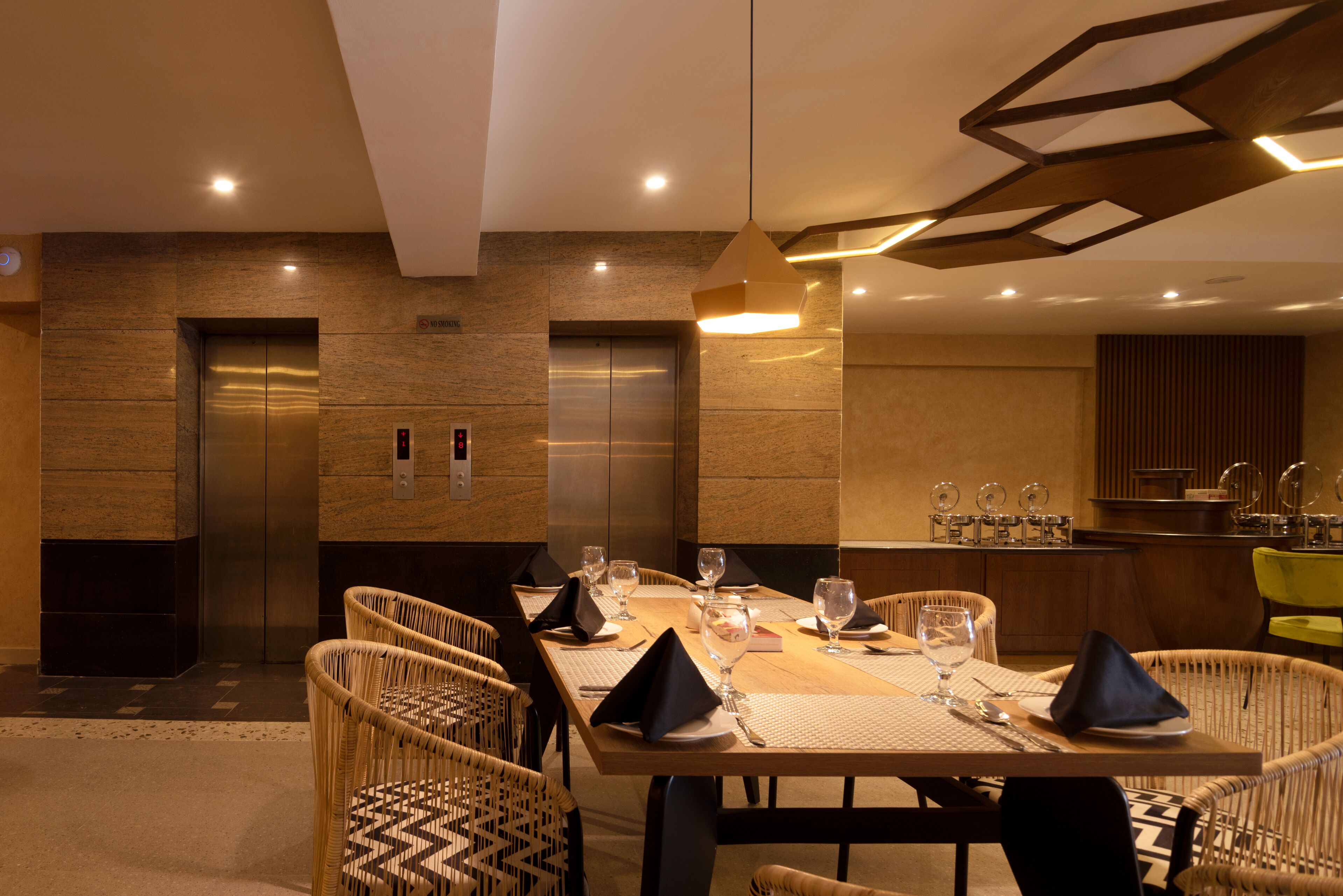 A dining area with a long table set for six, surrounded by wicker chairs, with two elevators in the background.