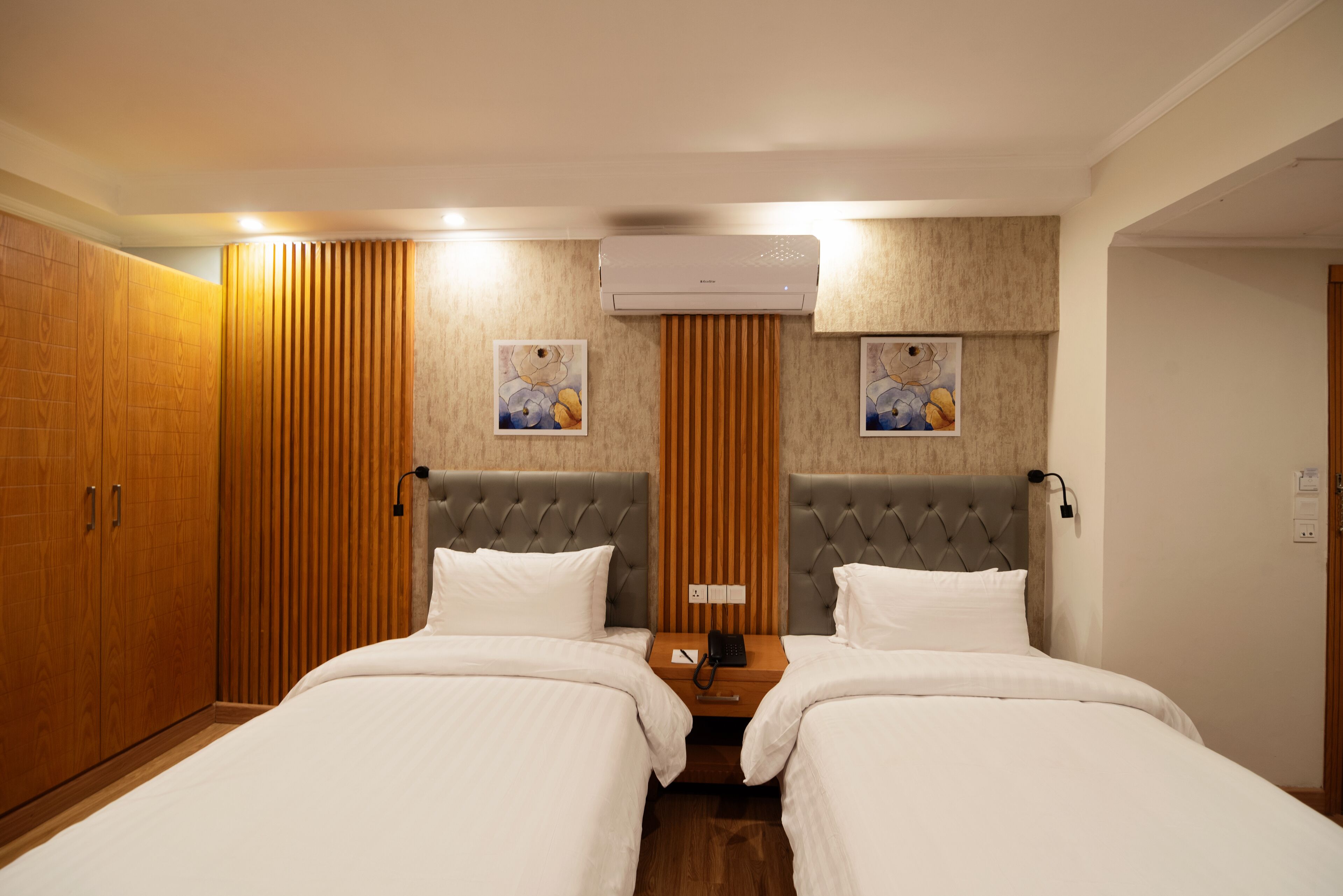 A hotel room with two beds, wooden paneling, a wall-mounted air conditioner, and artwork on the walls.