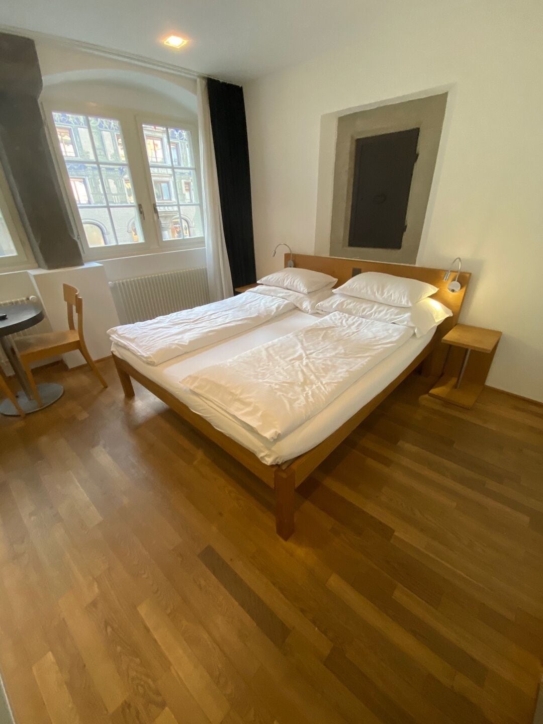 A bedroom with a double bed, a bedside table, a chair, a window with curtains, and a radiator.