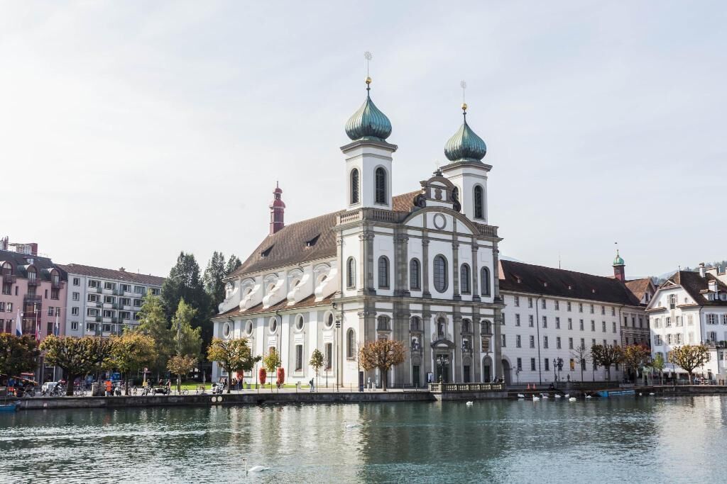 A historic building with two green domes and a central clock tower, located by a calm river with other buildings in the background.