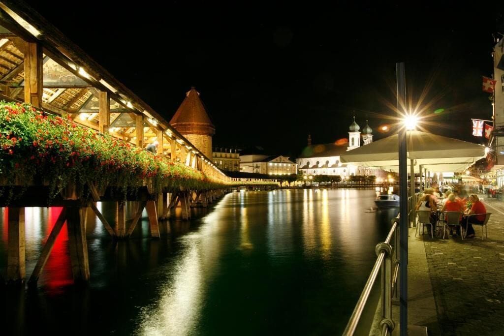 A riverside walkway with a wooden bridge, a tower, and a row of shops with outdoor seating.