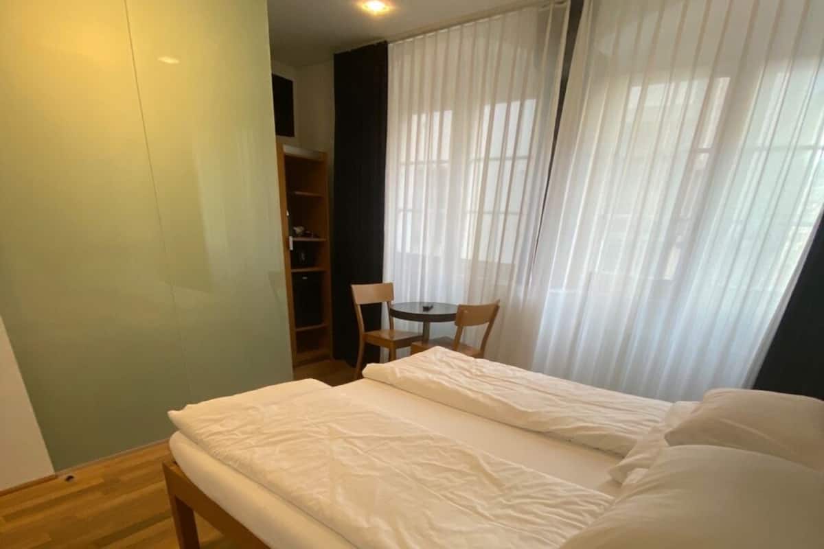 A bedroom with a large bed, wooden flooring, a wardrobe, a small table with chairs, and sheer curtains.